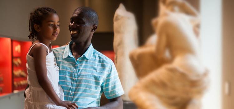 a father and his daughter smile and look at sculptures in an art museum